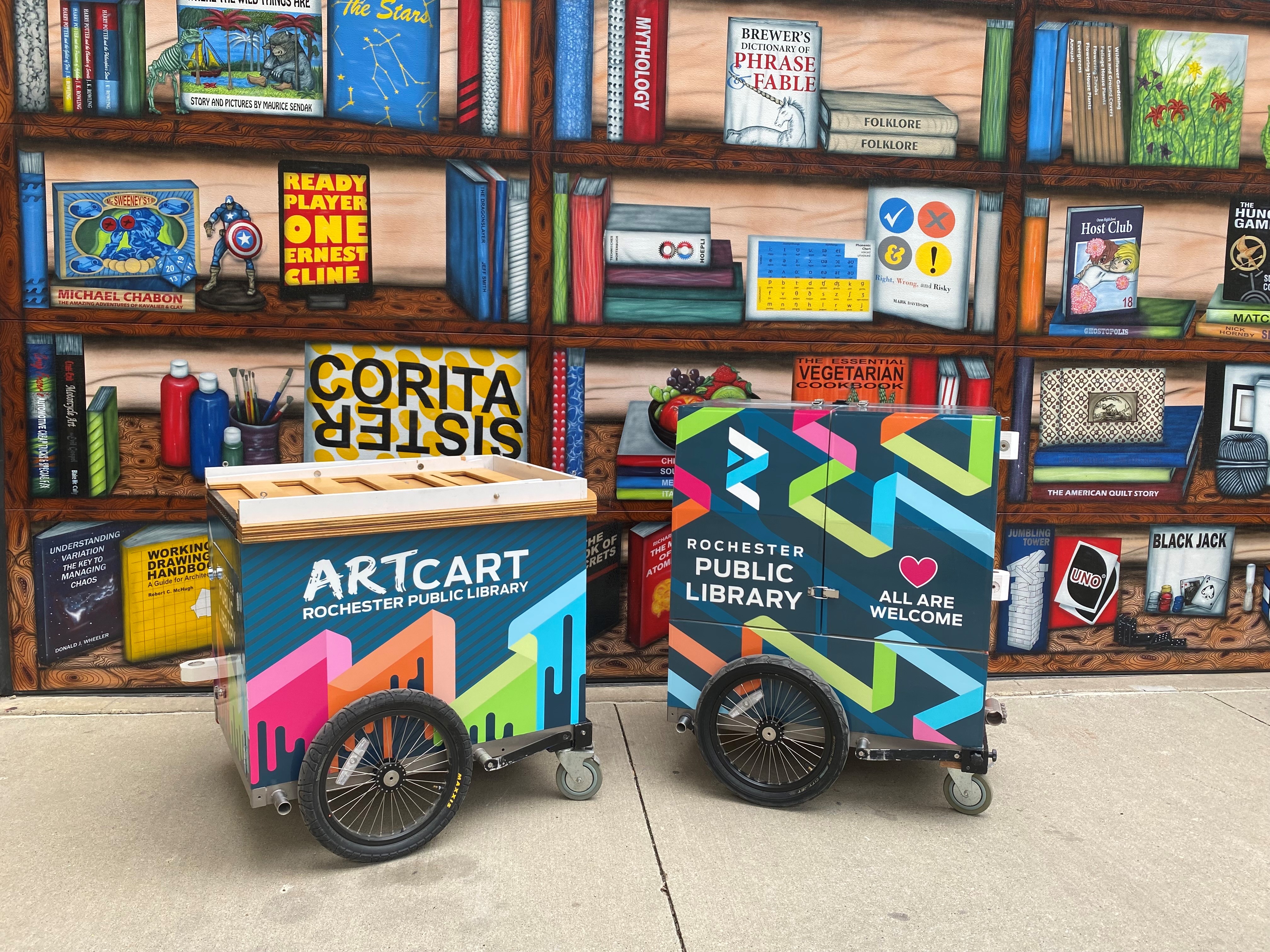 Photo of ArtCart and BookBike trailers in front of painted bookshelf mural