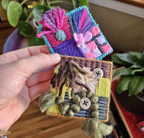 a hand holding two squares of plastic canvas decorated with yarn