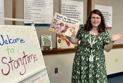 woman in green dress holding a book next to a "welcome to storytime" sign